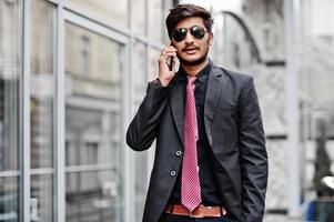 jonge Indiase man op pak, stropdas en zonnebril poseerde buiten, sprekend op de telefoon. foto