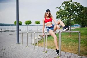 sexy krullend model meisje in rode top, jeans denim shorts, cup zonnebril en sneakers gesteld tegen stalen rails. foto