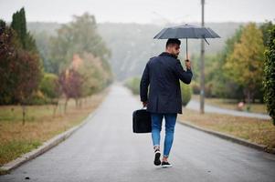 modieuze lange Arabische baard man draagt zwarte jas met paraplu en tas geval gesteld op regenweer dag. foto