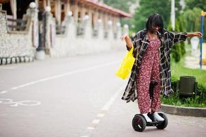 mooie afrikaanse amerikaanse vrouw die segway of hoverboard gebruikt. zwart meisje met gele stoffen eco-tassen recycling symbool. foto