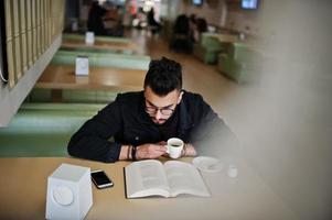 arabische man draagt een zwarte spijkerjas en een bril die in café zit, boek leest en koffie drinkt. stijlvolle en modieuze Arabische modelman. foto