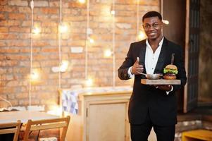 respectabele jonge afro-amerikaanse man in zwart pak houdt dienblad met dubbele hamburger vast en toont duim tegen bakstenen muur van restaurant met verlichting. foto