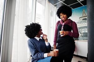 twee Afro-Amerikaanse vrouw met krullend haar draagt truien met kopjes thee in café binnen. foto