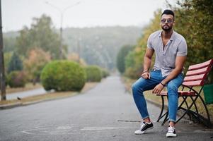 modieuze lange Arabische baard man slijtage op shirt, jeans en zonnebril gesteld op park op bankje. foto