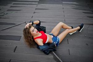 sexy krullend model meisje in rode top, jeans denim shorts, leren jas en sneakers op de vloer gesteld. foto