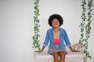 aantrekkelijke afro-amerikaanse vrouw met afro-haar op rok en jeansjasje, geposeerd in witte kamer op schommel. modieus zwart model. foto