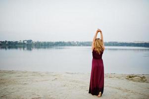 achterkant van blonde sensuele barefoot vrouw in rode marsala jurk poseren tegen meer. foto