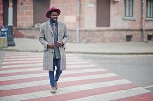 stijlvolle Afro-Amerikaanse man model in grijze jas, jas stropdas en rode hoed lopen op zebrapad. foto