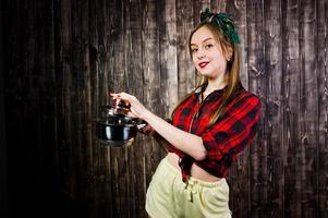 jonge grappige huisvrouw in geruit overhemd en gele korte broek pin-up stijl met steelpan op houten achtergrond. foto