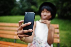 geweldige Afro-Amerikaanse modelvrouw in groene broek en zwarte hoed poseerde op een bankje in het park en maakte selfie op mobiele telefoon. foto