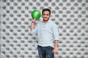 stijlvolle Aziatische man in jeans shirt staan met bowlingbal bij de hand tegen zilveren muur achtergrond. foto