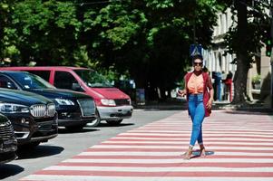 stijlvolle Afro-Amerikaanse vrouw lopen op zebrapad of zebrapad met mobiele telefoon bij de hand. foto