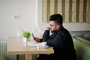 arabische man draagt een zwarte spijkerjas en een bril die in café zit, boek leest en koffie drinkt. stijlvolle en modieuze Arabische modelman. mobiele telefoon bij de hand. foto