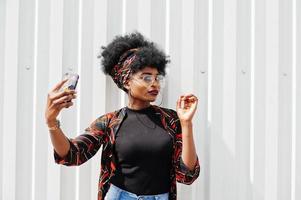 afrikaanse vrouw met afro-haar, in spijkerbroek en bril poseerde tegen een witte stalen muur en maakte een selfie via de telefoon. foto