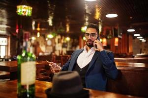 knappe goed geklede arabische man met glas whisky en sigaar houden mobiele telefoon, geposeerd in pub. foto