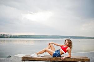 sexy krullend model meisje in rode top, jeans denim shorts, brillen en sneakers gesteld op bankje tegen meer. foto