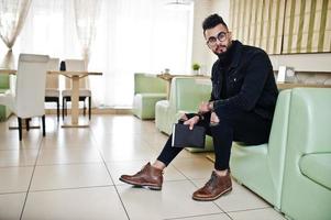 arabische man draagt een zwarte spijkerjas en een bril in café, lees boek. stijlvolle en modieuze Arabische modelman. foto