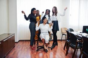 vijf multiraciale zakelijke dames staan op kantoor en rollen vrouw op stoel. diverse groep vrouwelijke werknemers in formele kleding die plezier hebben. foto