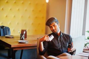 zelfverzekerde jonge indiase man in zwart shirt zit in café en laat zijn creditcard zien. foto