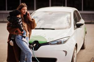 jonge moeder met kind elektrische auto opladen bij het elektrische tankstation en spreken op mobiele telefoon. foto