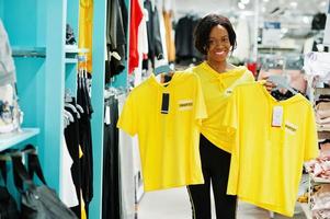 Afro-Amerikaanse vrouwen in trainingspakken winkelen bij sportkleding winkelcentrum tegen planken. ze koos geel t-shirt. sport winkel thema. foto