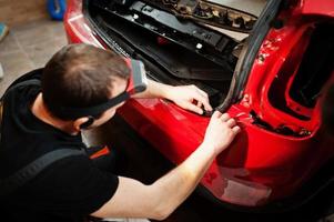 autoservicemedewerker plaatste anti-grindfilm op een rode carrosserie in de detailleringsvoertuigwerkplaats. autobescherming met speciale folies. foto