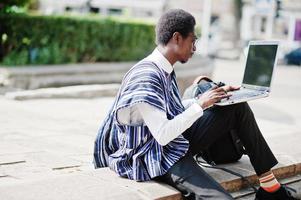 Afrikaanse man student in traditionele kleding en bril met rugzak, werken op laptop buiten. foto