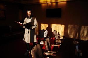 knappe Afro-Amerikaanse man poseren in kamer met zonlicht schaduwen in zwarte hoed en beige jas. foto