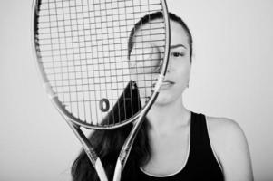 zwart-wit portret van een mooie jonge vrouw in sportkleding die een tennisracket vasthoudt terwijl hij tegen een witte achtergrond staat. foto