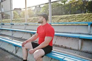 jonge brute bebaarde gespierde man draagt een rood shirt, korte broek en pet in het stadion. foto