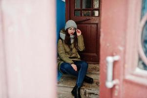 portret van brunette meisje in grijze sjaal en muts, bril tegen ingang. foto