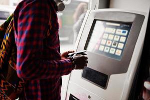 Afro-Amerikaanse man in geruit overhemd met rugzak portemonnee in de hand houden tegen ATM. zwarte man reiziger. foto