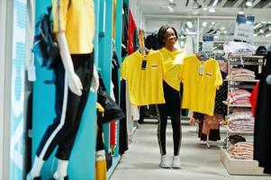 Afro-Amerikaanse vrouwen in trainingspakken winkelen bij sportkleding winkelcentrum tegen planken. ze koos geel t-shirt. sport winkel thema. foto