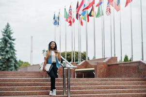 afrikaanse student vrouw poseerde met rugzak en schoolspullen op het erf van de universiteit, tegen vlaggen van verschillende landen. foto