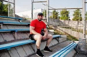 jonge brute bebaarde gespierde man draagt een rood shirt, korte broek en pet in het stadion. foto