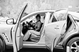 rijke zakelijke afrikaanse vrouw zit op de bestuurdersstoel bij zilveren suv-auto met geopende deur. foto
