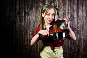 jonge grappige huisvrouw in geruit overhemd en gele korte broek pin-up stijl met steelpan op houten achtergrond. foto