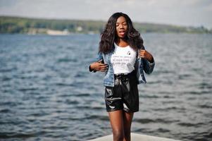 Afro-Amerikaanse donkere huid slank model poseerde in een zwarte broek en jeans jasje tegen de zee. foto