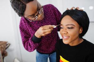 Afro-Amerikaanse vrouw die make-up toepast door visagist bij beauty salon. foto