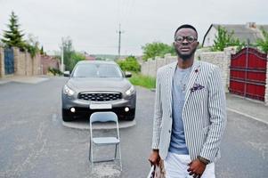 rijke en stijlvolle Afro-Amerikaanse man in blazer en witte broek, bril las tijdschrift tegen zijn suv-auto. foto