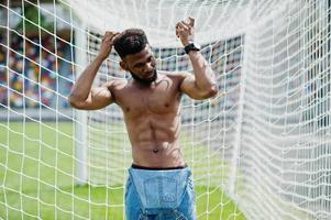 knappe sexy Afro-Amerikaanse gespierde blote torso man op jeans overall gesteld op groen gras tegen voetbal poorten van stadion veld. modieuze zwarte man portret. foto