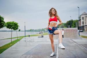 sexy krullend model meisje in rode top, jeans denim shorts, brillen en sneakers gesteld op skatepark. foto