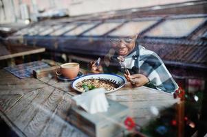 afrikaanse vrouw in geruite cape en bril die in café zit en dessert eet. foto