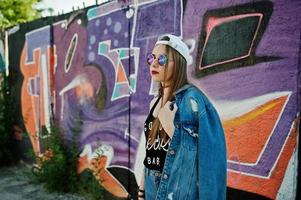 stijlvolle casual hipster meisje in pet, zonnebril en spijkerbroek dragen, muziek luisteren van koptelefoon van mobiele telefoon tegen grote graffiti muur. foto