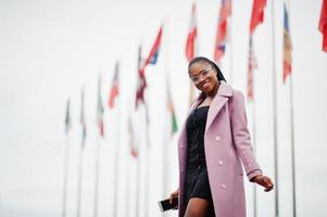 jonge stijlvolle mooie afro-amerikaanse vrouw in straat, gekleed in een mode-outfitjas, tegen vlaggen van verschillende landen van de wereld, met mobiele telefoon bij de hand. foto