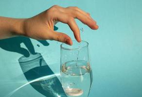 tegen een blauwe achtergrond laat een hand een oplossende koolzuurhoudende aspirinetablet in een glas water vallen foto