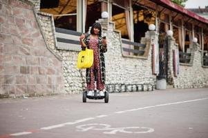 mooie afrikaanse amerikaanse vrouw die segway of hoverboard en mobiele telefoon gebruikt. zwart meisje met gele stoffen eco-tassen recycling symbool. foto