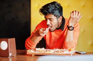 zelfverzekerde jonge indiase man in oranje trui zit bij pizzeria en eet pizza. foto