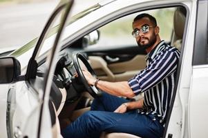 succesvolle arabische man draagt een gestreept shirt en een zonnebril poseert achter het stuur van zijn witte suv-auto. stijlvolle arabische mannen in transport. foto