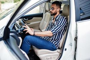 succesvolle arabische man draagt een gestreept shirt en een zonnebril poseert achter het stuur van zijn witte suv-auto. stijlvolle arabische mannen in transport. foto